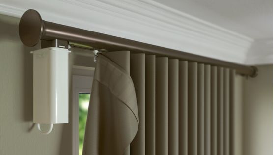 Motorised curtain track with visible motor unit mounted at the end of a window, showing the curtain partially wrapped around the track beneath a metal curtain pole.