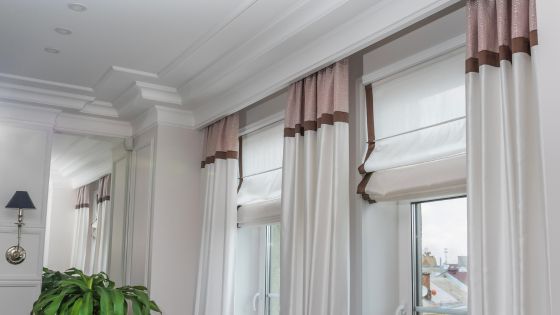lovely white room with 2 large long windows that have blinds in them and them ad then a long curtain pole which spans across the whole wall with 3 curtains hanging.